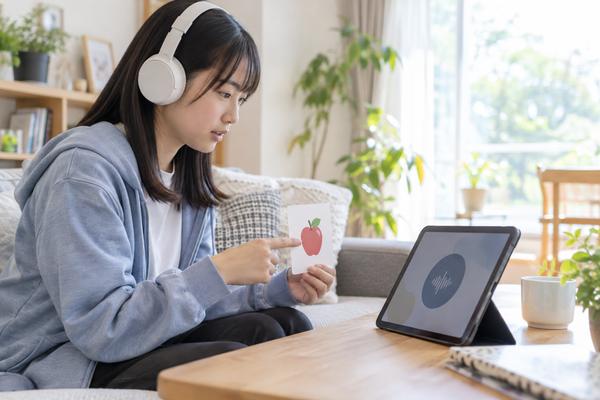 ヘッドホンを付けた中学生が自宅でタブレットの英語音声を聞きながら、声に出して確認している様子