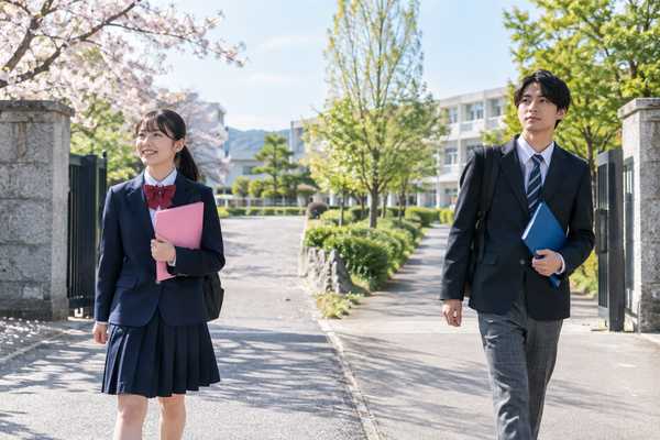 桜の咲く校門前の通学路を、中学生と高校生がそれぞれの制服姿で歩いている様子