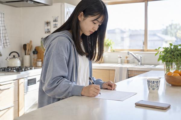 朝のキッチンカウンターで、スマートフォンを伏せた私服の女子生徒が一枚の問題用紙に向かって解き直している様子