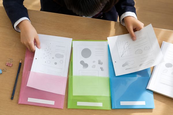 制服の中学生が返却された答案やレポートを、ピンク・緑・青の3色のファイルに観点ごとに分けて入れている手元の俯瞰
