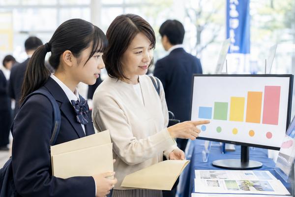 進路相談会で同じ目盛りのグラフを見ながら候補校を比べる制服の高校生と保護者