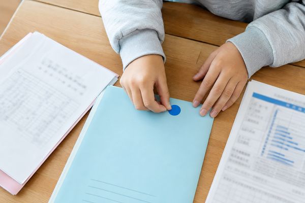 中学生が机の上で通知表と返却物を見ながら、一教科分のファイルに青い丸シールを貼っている手元
