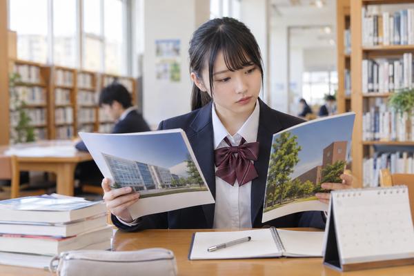 制服姿の高校生が高校の図書館で、二つの大学の入試情報の冊子を見比べ、カレンダーで期間を確かめている様子