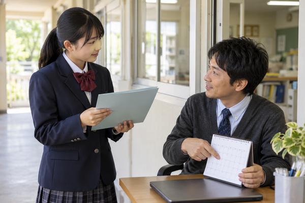 職員室の窓際で、制服姿の中学生が資料を手に、先生からカレンダーで次の評価機会を示されながら質問している様子