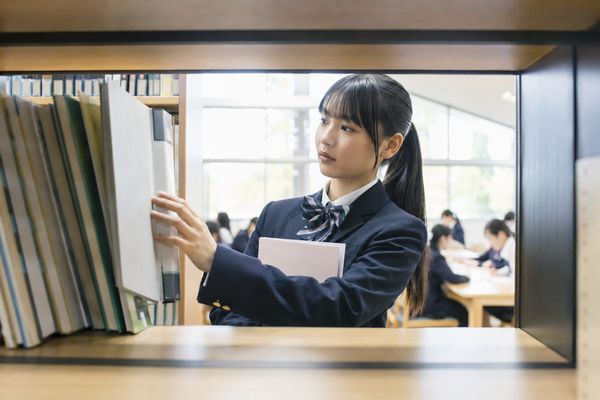 学校図書室の書架で、制服姿の女子高校生が資料のバインダーを棚から取り出している様子