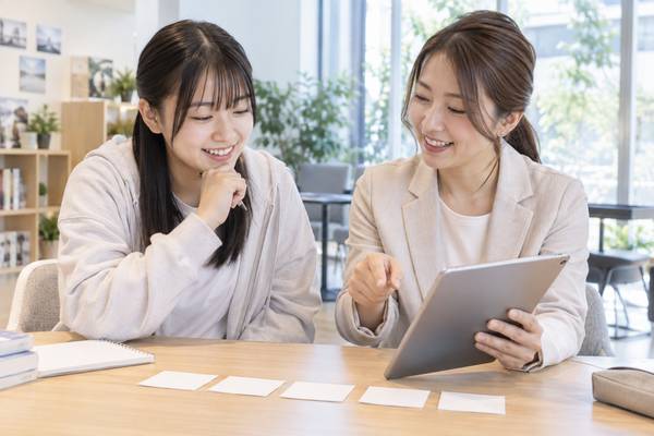 私服の高校生と女性講師が明るい相談スペースでタブレットを見ながら、教科書から過去問へ進む学習の順番を確認している様子