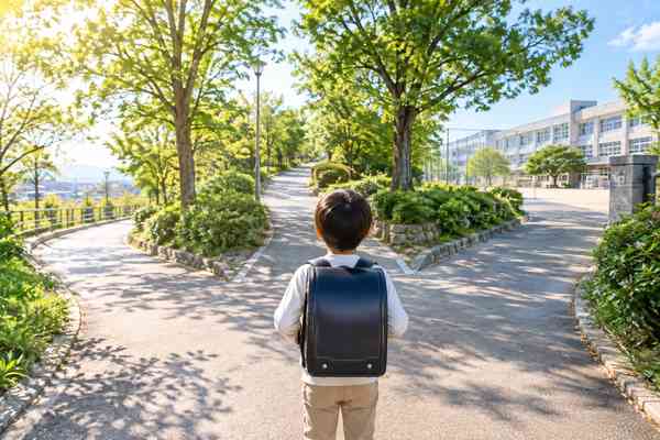 ランドセルを背負った小学生が、学校近くの分かれ道で行き先を考えて立ち止まっている様子