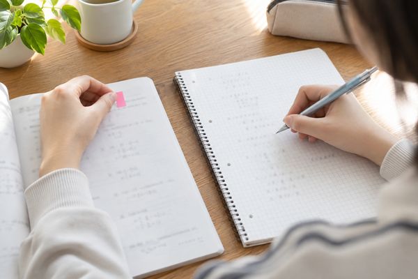 自宅の机で、私服の中学生が間違えた一問にピンクの付箋を貼り、ノートに解き直している手元のアップ。全問ではなく一問から復習する様子
