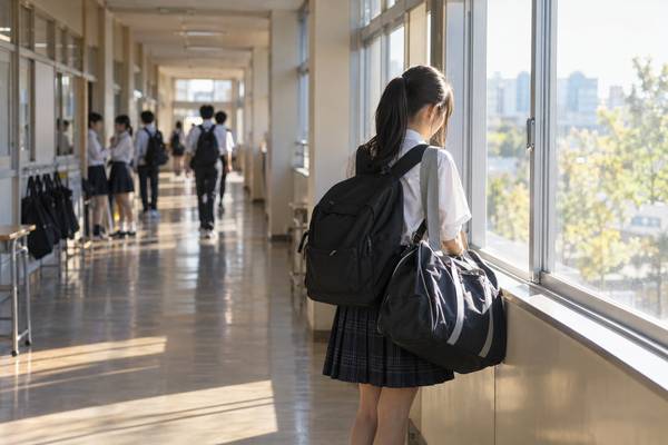 放課後の明るい校舎の廊下で、大きな部活のバッグとリュックを抱えた生徒が窓際で足を止めている様子