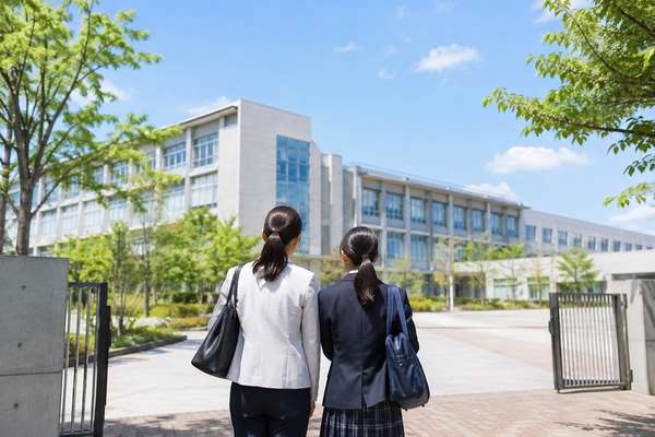 制服姿の中学生と保護者が学校見学で高校の校舎を並んで見上げている後ろ姿