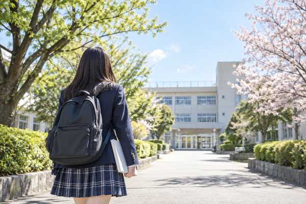 桜の咲く並木道を通り、英語の問題集を持って学校へ歩いていく制服姿の生徒の後ろ姿