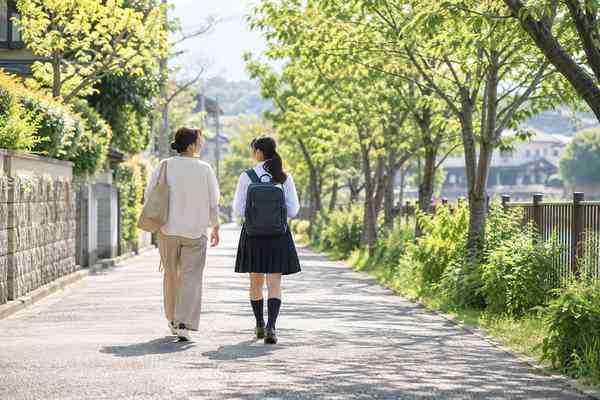 緑の多い帰り道を、制服姿の生徒と母親が並んで歩きながら話している後ろ姿