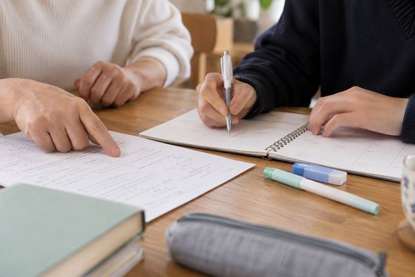 ダイニングテーブルで大人の手が答案の一点を指差し、生徒の手がペンで週間予定表に次の一手を書き込んでいる手元