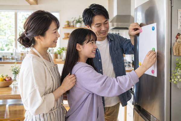 冷蔵庫に身長の記録用紙をマグネットで貼りながら笑顔で話す家族3人