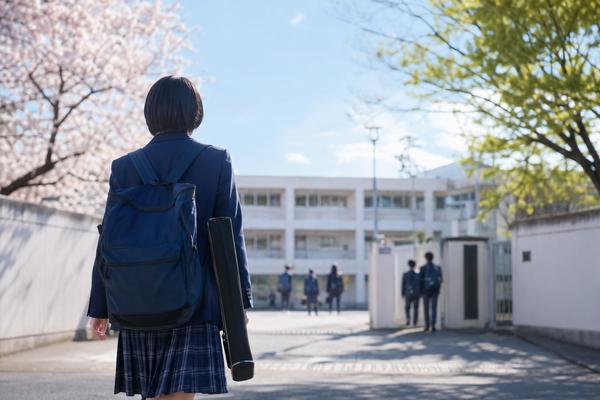 画材ケースとリュックを持ち、朝の明るい校門へ歩いて向かう中学生の後ろ姿