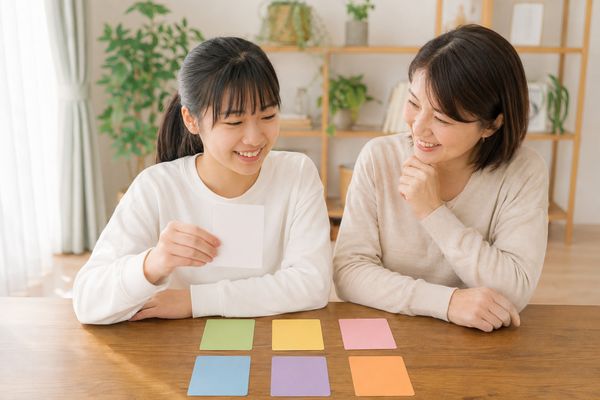 自宅のテーブルに並べた色違いのカードから、次の目標を一枚だけ選ぶ中学生と見守る保護者