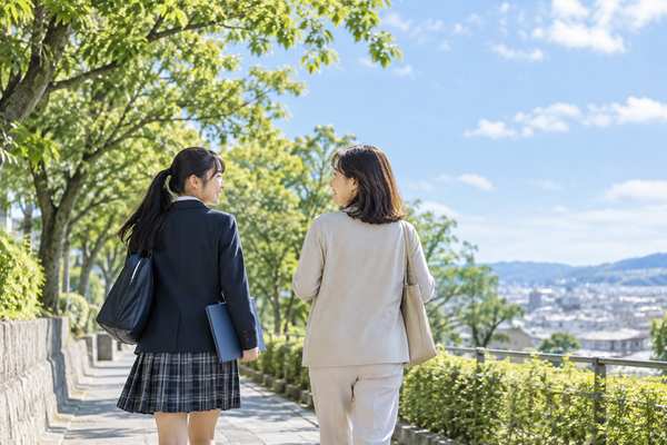 通学路を並んで歩きながら、母親と高校生が笑顔で話している場面