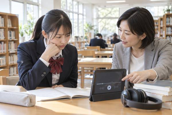 学校の図書室で、リスニング音声を前に困った表情の高校生と原因を指し示す先生