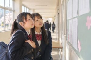 春の高校の廊下で、掲示板に貼られた新しいクラス分け表を見上げる制服姿の高校生2人