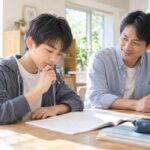 明るいダイニングで宿題の問題に手を止めて考える中学生と、そばで静かに見守る父親