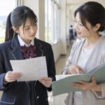 高校の廊下で答案を手にした生徒が先生に成績を相談している