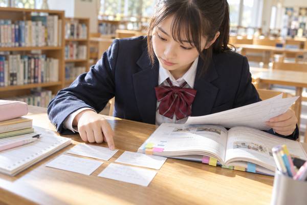 学校の図書室で英語のテスト範囲表と教科書を見比べる中学一年生