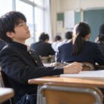授業中に手元のノートから視線を外している制服の中学生と、黒板の前で説明を続ける先生