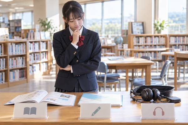 学校図書室で、本・鉛筆・ヘッドホンのアイコンが付いた3種類の英語テスト教材を見比べる制服の中学生
