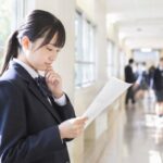 明るい校舎の廊下で、返却された定期テストの答案を手に持ち、成績を考え込む制服の中学生