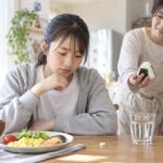 朝食の並んだ食卓で、食欲の出ない中学生におにぎりを差し出す保護者と、机に置かれた一粒のブドウ糖