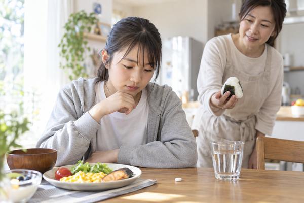 朝食の並んだ食卓で、食欲の出ない中学生におにぎりを差し出す保護者と、机に置かれた一粒のブドウ糖