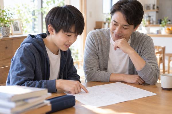 日当たりのよいダイニングで、中学生の息子と父親が返却された5教科のテストを一緒に見ながら笑顔で話している様子