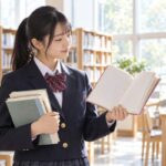 明るい図書室で何冊もの本を抱え、開いた一冊を見比べている制服姿の高校生