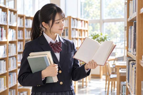 明るい図書室で何冊もの本を抱え、開いた一冊を見比べている制服姿の高校生