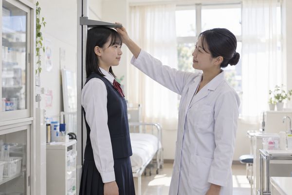 学校の保健室で、制服姿の中学生が身長計の前に立ち、養護教諭が測定バーを頭の高さに合わせている様子