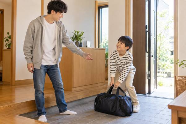 自宅の玄関で、兄が弟に鞄を持つよう頼み、弟が鞄を持ち上げている場面