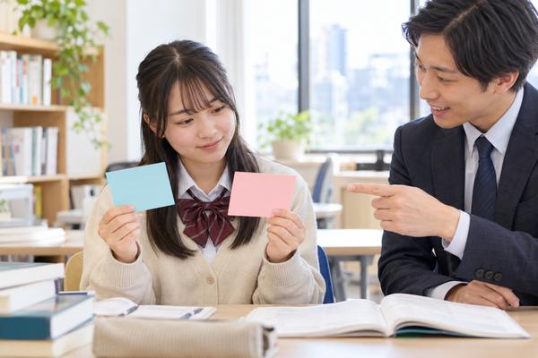 二枚のカードを手に持ち、はい・いいえ型と疑問詞型の違いを先生と確認している制服姿の高校生