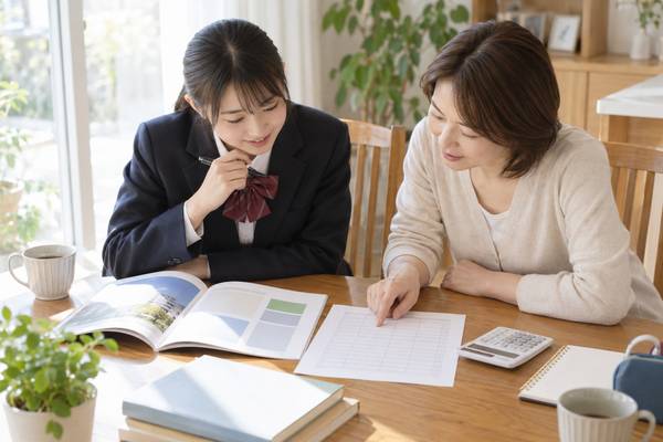 ダイニングテーブルで大学案内と得点表と電卓を並べ、換算後の得点率を保護者と一緒に確かめる制服姿の高校生