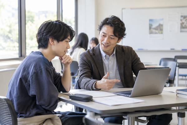 大学の教室でノートパソコンのシラバスを見ながら教員に相談する私服の学生
