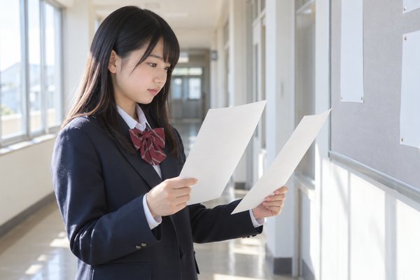 制服姿の女子中学生が、明るい校舎の廊下で2枚の配布プリントを見比べている様子