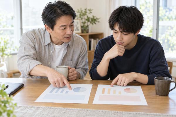 明るいダイニングで、父親と浪人生の息子が2種類の調査結果を並べ、どこが違うのかを指さして話し合っている様子