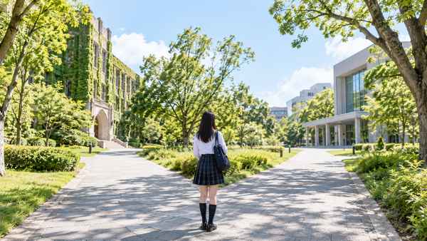 大学キャンパスの分かれ道に立ち、校舎の異なる2つの進路を見比べる制服姿の高校生