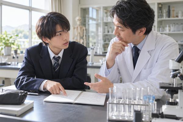 理科室でノートの一か所を指さし、分からない場所を先生に質問する高校生