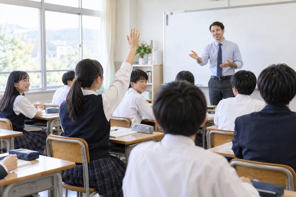 英語の授業で手を挙げて答えようとする中学生と、教室の前で問いかける先生