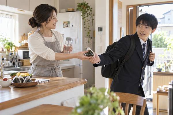 登校前の玄関で、おにぎりと水筒を手渡す保護者と、制服姿で受け取る中学生