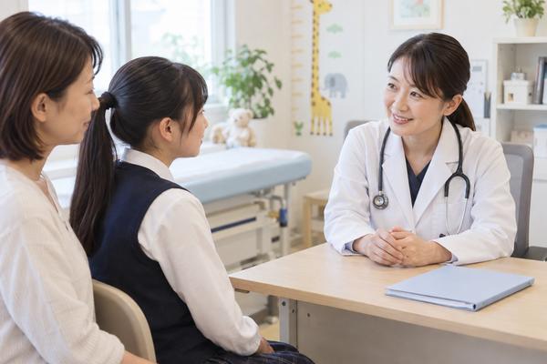 明るい小児科の診察室で、保護者と中学生が医師と向かい合い、成長の記録を見ながら穏やかに相談している様子