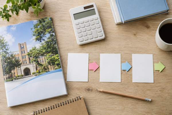 机の上に大学案内と電卓、3枚の白いカードと矢印を並べ、目標得点率を決める3つの手順を表した俯瞰写真