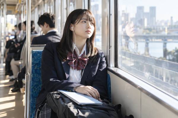 朝の通学電車でイヤホンを着けたまま、教材を開かずに窓の外を眺める制服姿の高校生