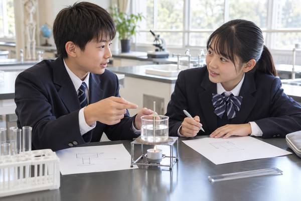 明るい理科室で、実験装置を前に図を指さしながら説明し合う制服姿の中学生2人