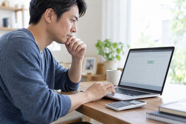 ノートパソコンの表計算画面に入れた合計の数式を、電卓を横に置いて二重に確かめている保護者の写真